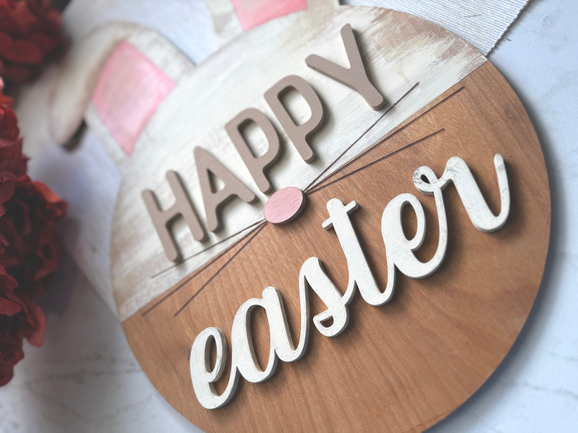 “Happy Easter” sign featuring a round base with bunny ears and a pink nose. The upper portion is distressed white with pink ears. The bottom is a stained wood.