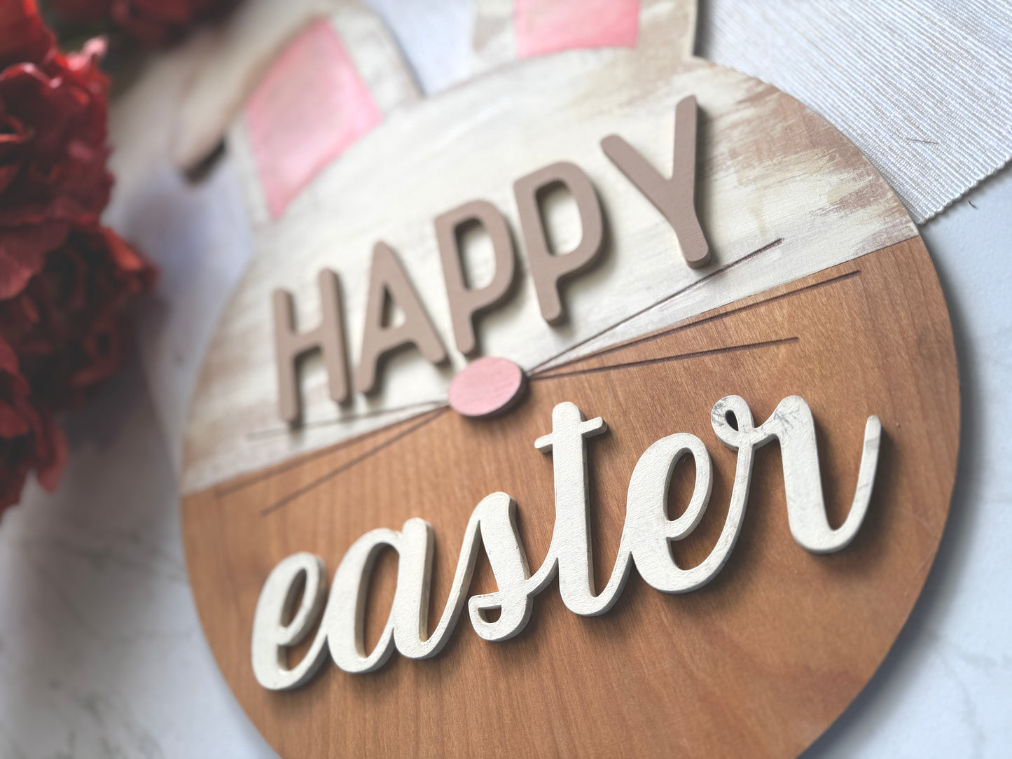 “Happy Easter” sign featuring a round base with bunny ears and a pink nose. The upper portion is distressed white with pink ears. The bottom is a stained wood.
