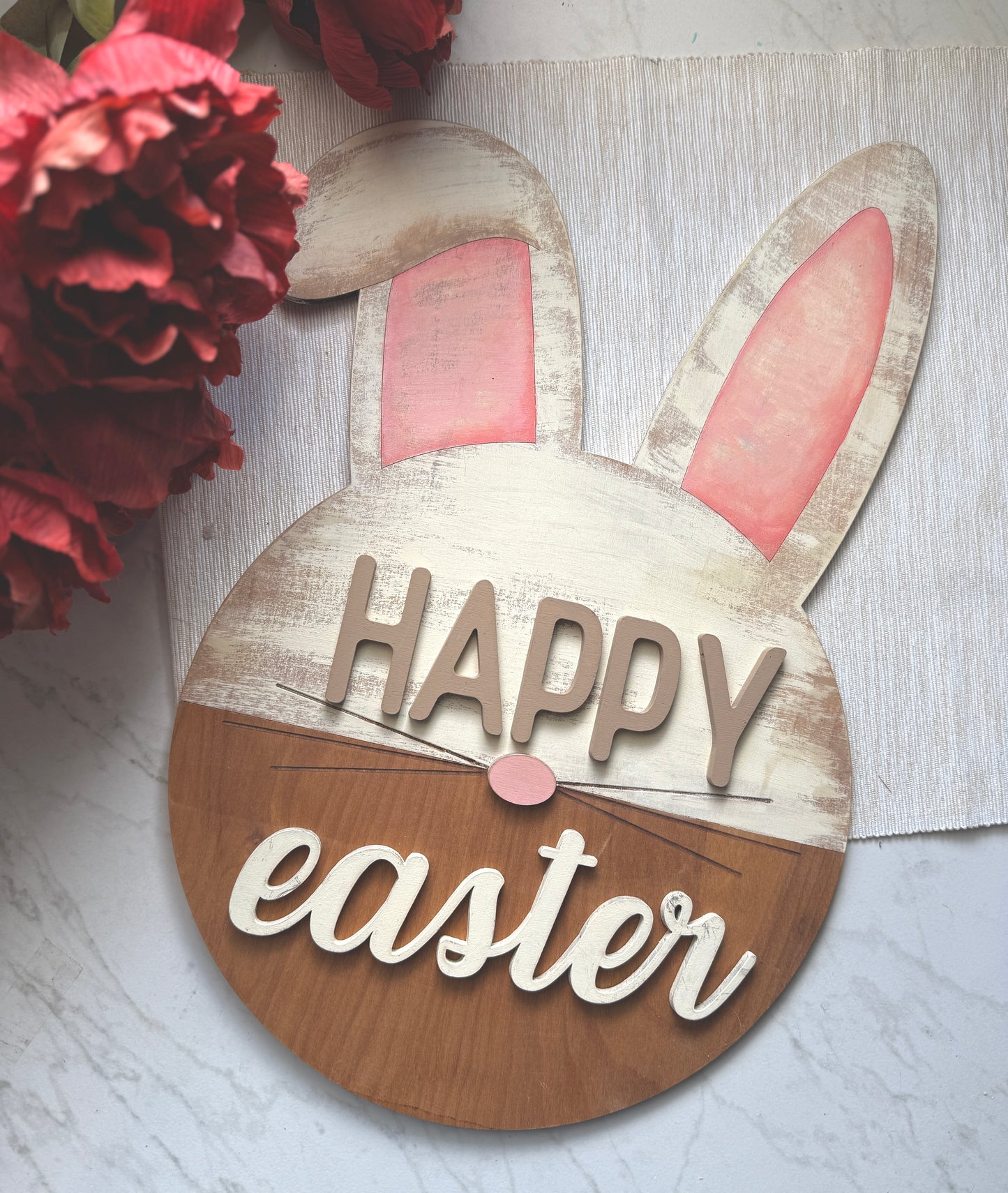 “Happy Easter” sign featuring a round base with bunny ears and a pink nose. The upper portion is distressed white with pink ears. The bottom is a stained wood.