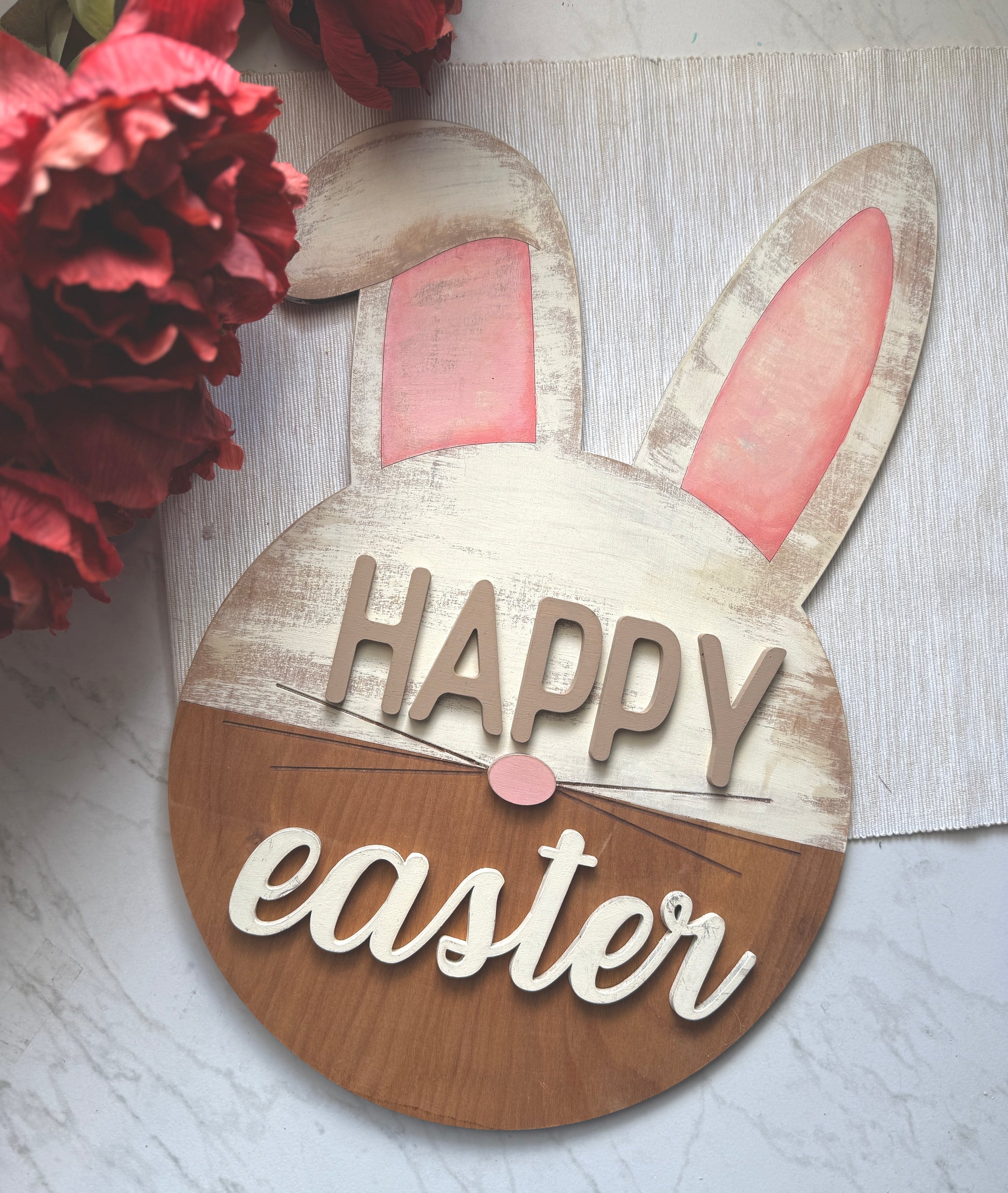 “Happy Easter” sign featuring a round base with bunny ears and a pink nose. The upper portion is distressed white with pink ears. The bottom is a stained wood.