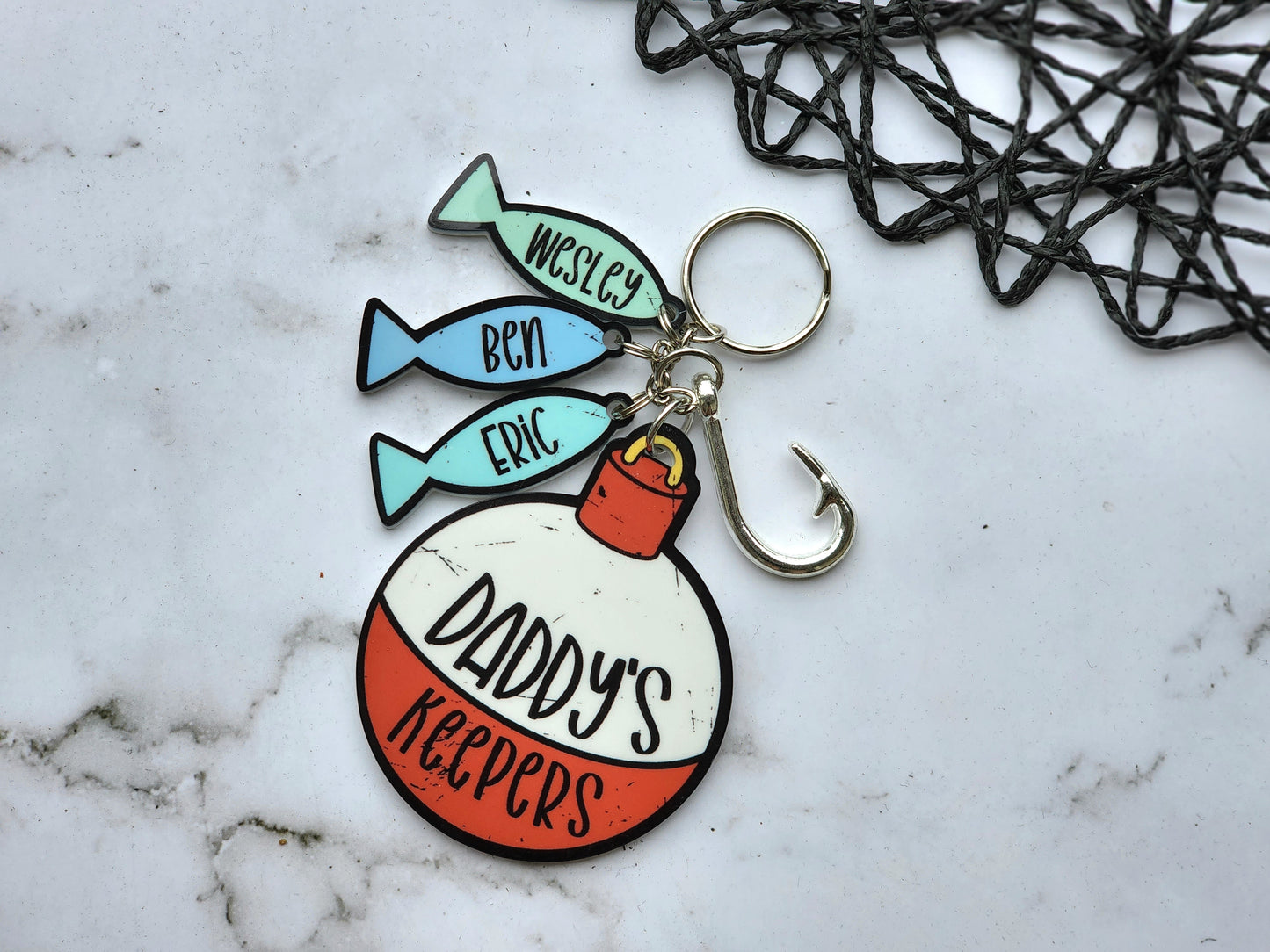 A cute little keychain made of acrylic featuring a bobber design reading "Daddy's Keepers" and 3 little fish reading eric, ben and wesley. The keychain also includes a faux fishing hook .