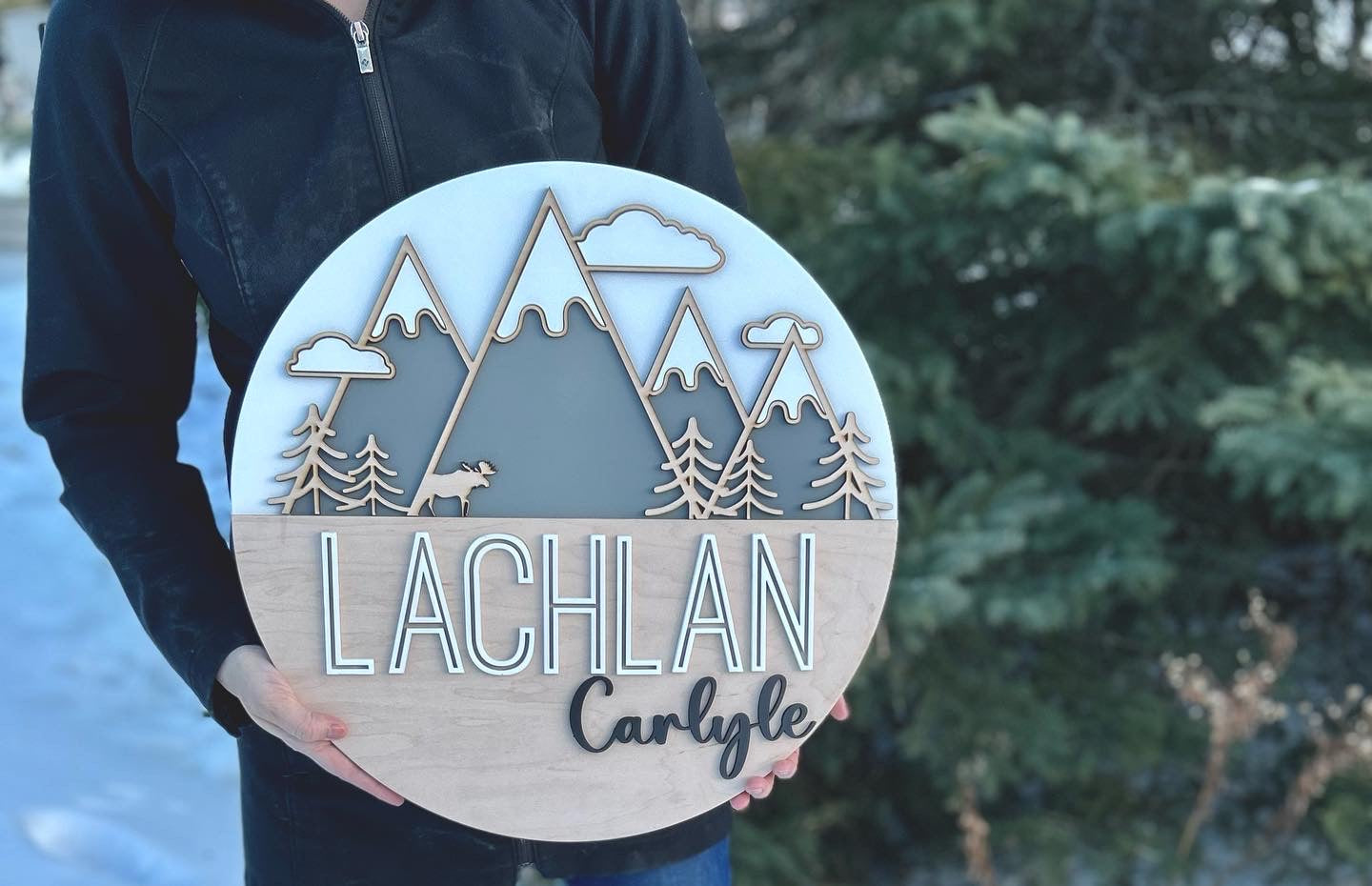 A white coloured round sign with 4 grey mountains with snowy tops and 3 clouds around the peaks. At the base of the mountains there are trees and a moose.   The bottom half of the sign is a maple backer, sanded beautifully featuring the name "Lachlan" in a bold, yet modern print painted white. to the bottom right in a smaller cursive font it reads "Carlyle"