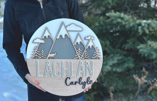 A white coloured round sign with 4 grey mountains with snowy tops and 3 clouds around the peaks. At the base of the mountains there are trees and a moose.   The bottom half of the sign is a maple backer, sanded beautifully featuring the name "Lachlan" in a bold, yet modern print painted white. to the bottom right in a smaller cursive font it reads "Carlyle"