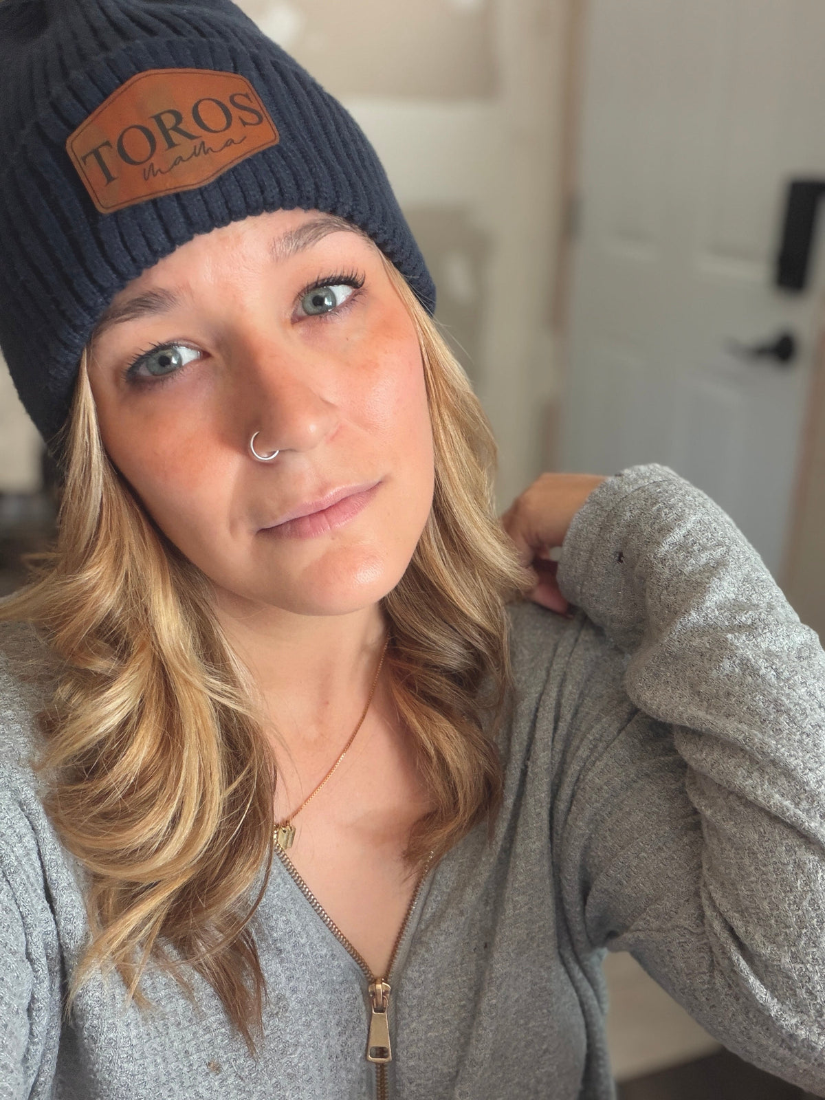 A woman wearing a dark navy ribbed knit beanie with a brown leather patch that reads 'TOROS' and a gray zip-up hoodie, photographed in an indoor setting.