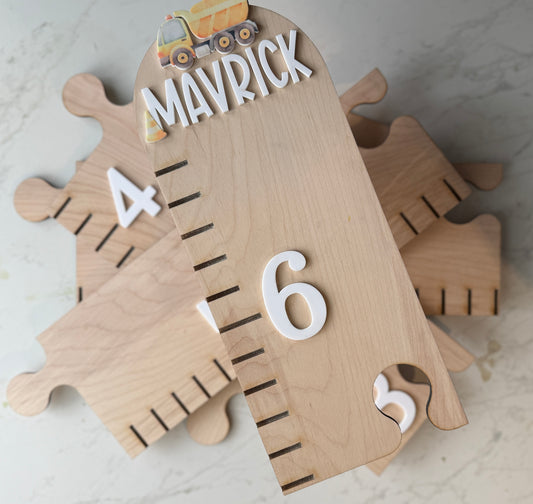 Wooden growth chart with puzzle pieces and numbers on a light background featuring a dump truck and the name ‘mavrick’