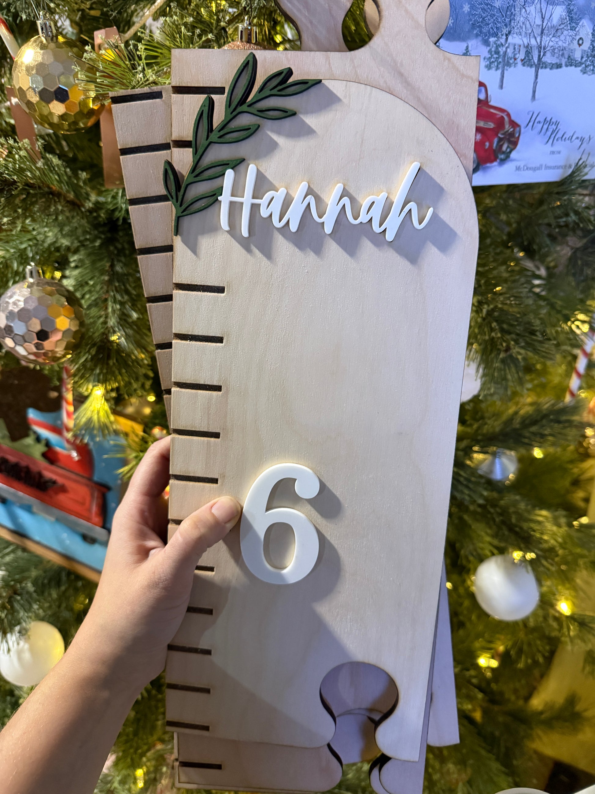 Wooden growth chart with 'Hannah' and number ‘6' held in front of a decorated Christmas tree.