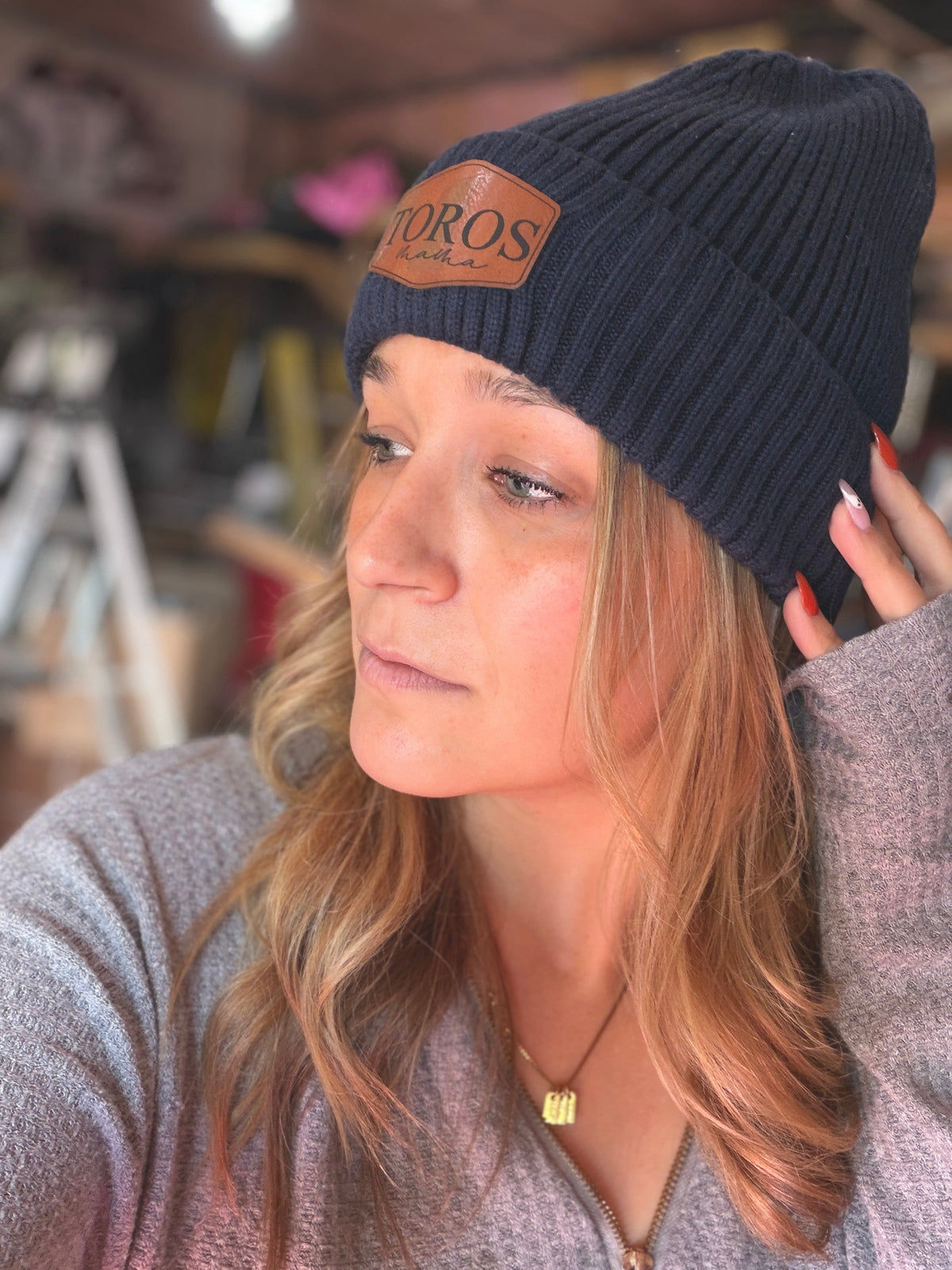 A woman wearing a dark navy blue ribbed knit beanie with a brown leather patch on the front that reads 'OROS'. She is wearing a gray knit top in a warm, slightly blurred indoor setting.